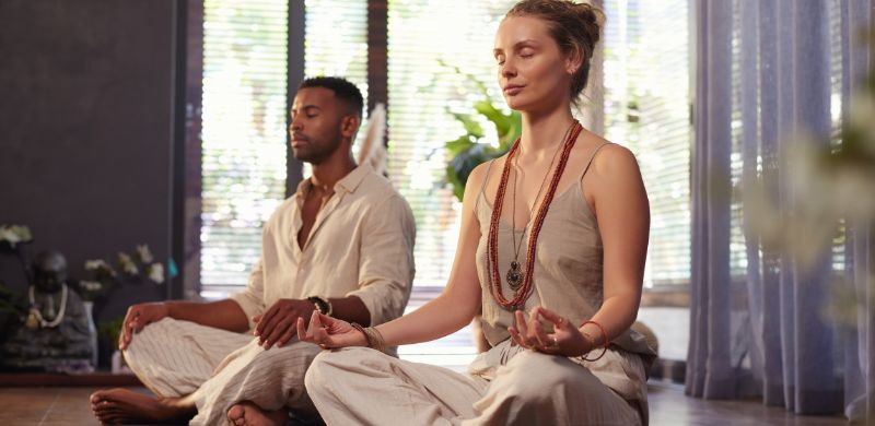 A man and a woman meditating in a calm, well-lit space, sitting on the floor with their eyes closed, focused and at peace.