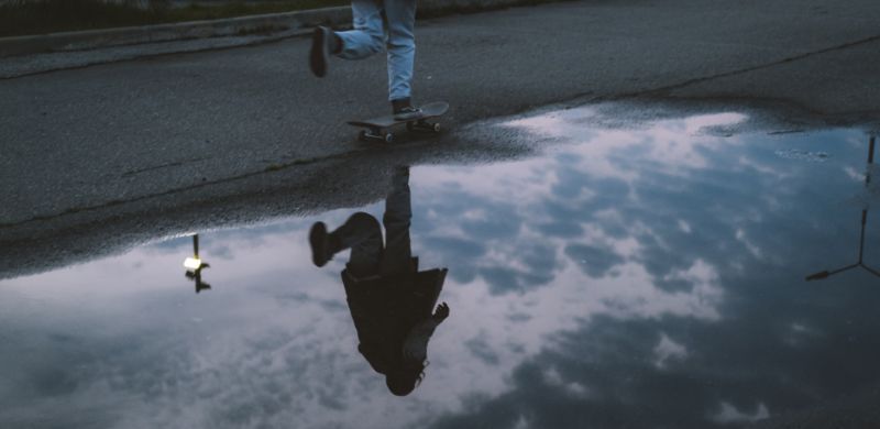 A person skateboarding on asphalt with their reflection visible in a puddle of water on the street below.